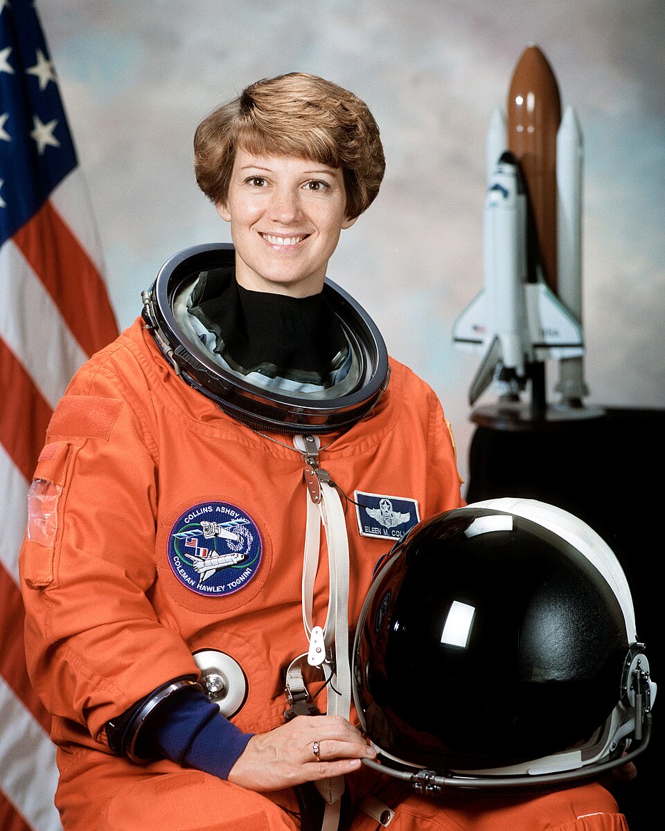 Eileen Collins in an orange flight suit beside a Space Shuttle model