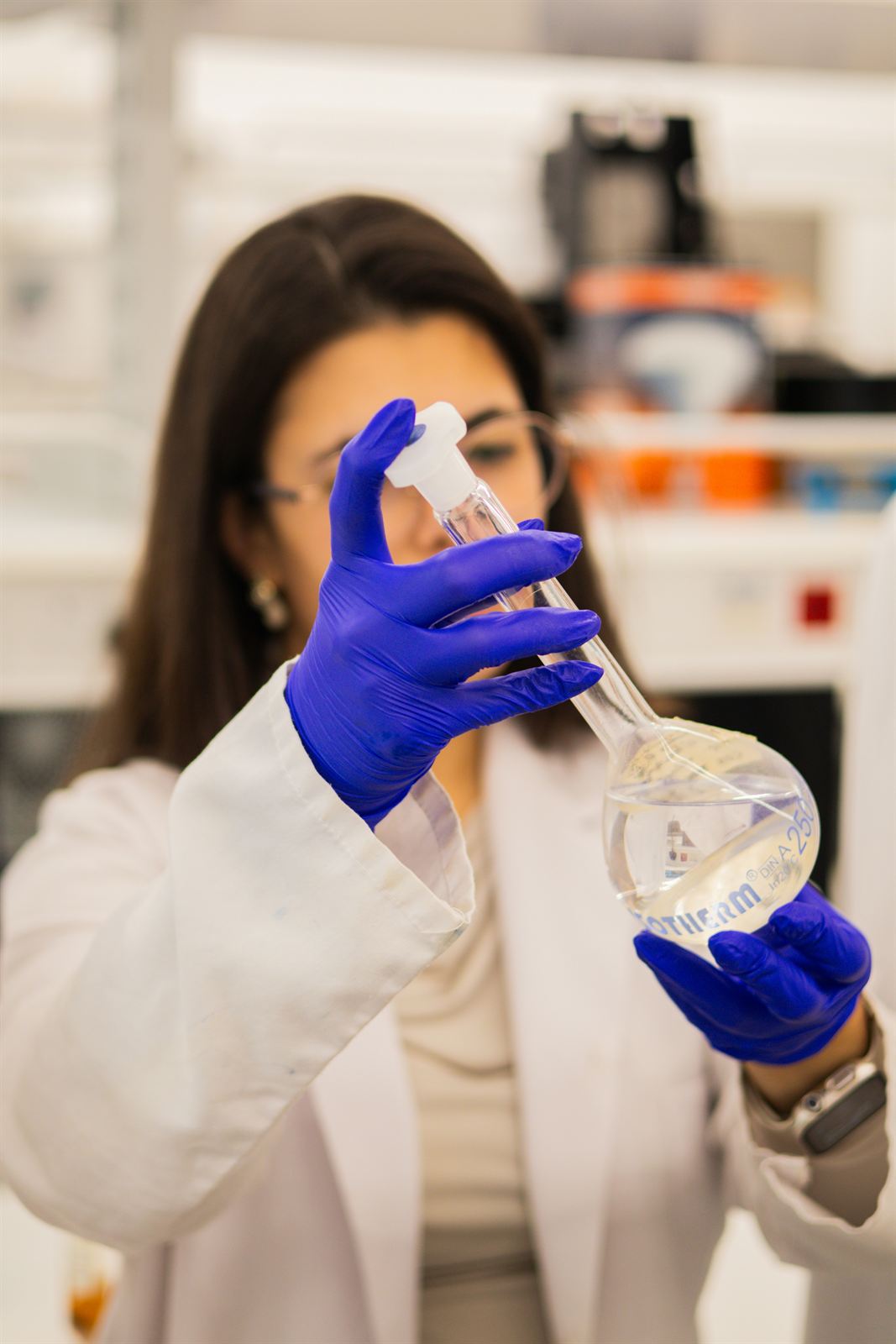 A researcher in a lab coat and blue gloves examining a flask