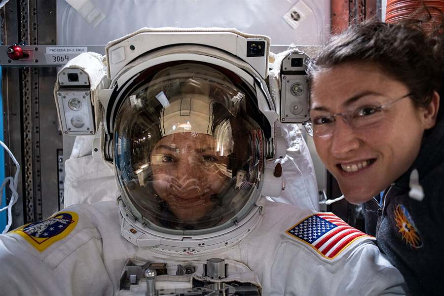 Two astronauts aboard the International Space Station, one wearing a spacesuit helmet, smiling at the camera