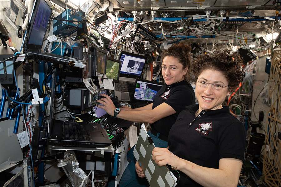Two women astronauts working at a crowded research workstation on the International Space Station